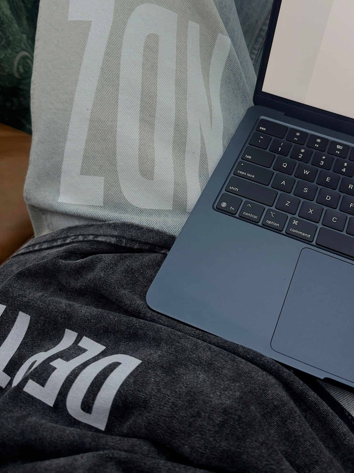 A dark slate blue laptop is open on a person's lap, showing a website on the screen. The person is wearing a light gray sweatshirt with large white letters and a dark gray sweatshirt underneath.
